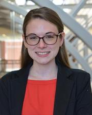 Ashley Taconet, Postdoctoral Researcher: a woman with long brown hair, glasses, and a black blazer over a scarlet shirt